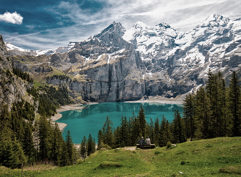 Un lac alpin bleu-vert avec des sommes enneigés en fond et une prairie alpestre avec des sapins devant.