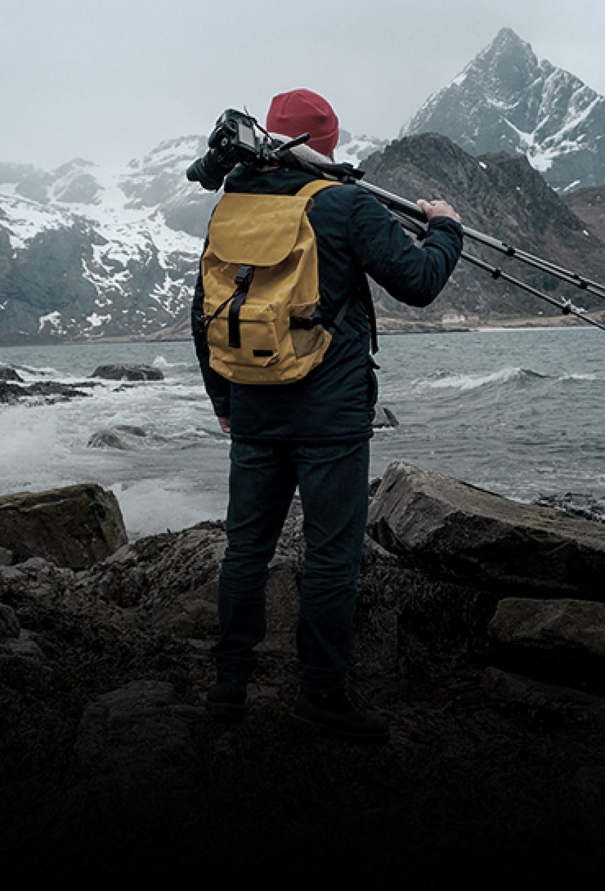 Homme dans les Alpes avec un bonnet rouge, un sac à dos jaune et un appareil-photo avec trépied sur l'épaule.